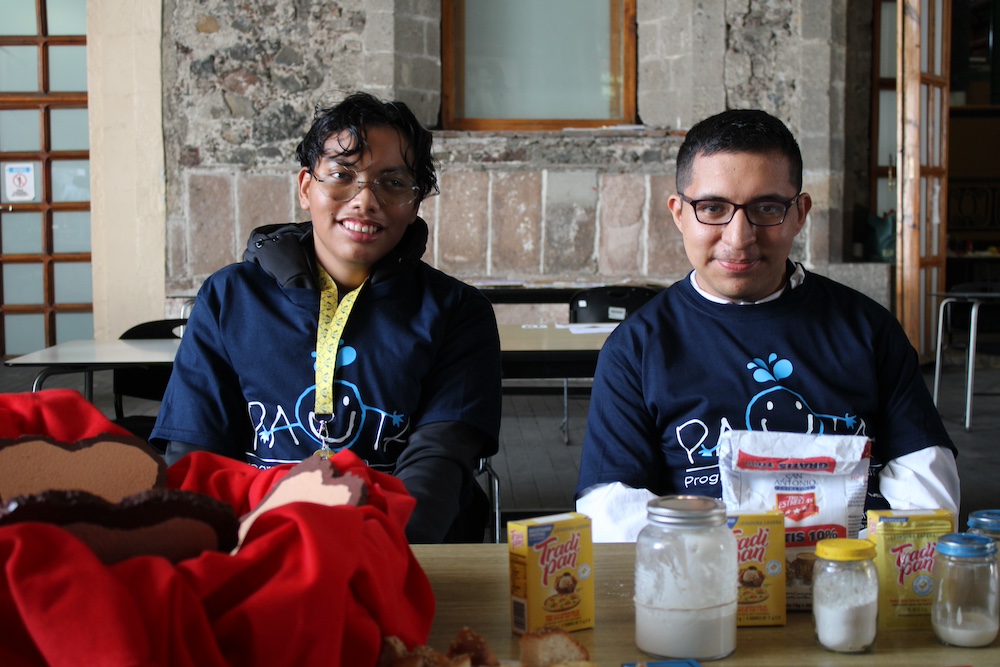 Dos beneficiarios de APAC que participan en el Club de Ciencias presentan su proyecto durante la Feria Estatal PAUTA 2026, con productos de cocina sobre la mesa que forman parte de su experimento.