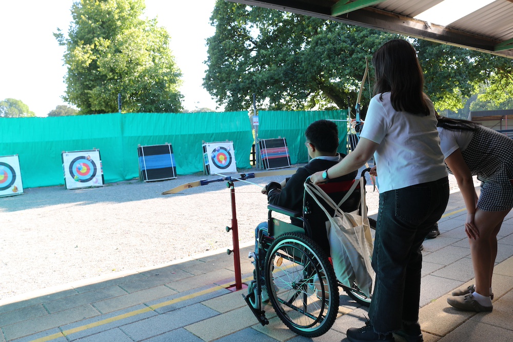 Una persona usuaria de silla de ruedas participa en una actividad de tiro con arco con apoyo de dos personas durante la Cumbre iCAN 2026 en Barretstown, Irlanda. La actividad forma parte de las experiencias recreativas desarrolladas durante el encuentro.