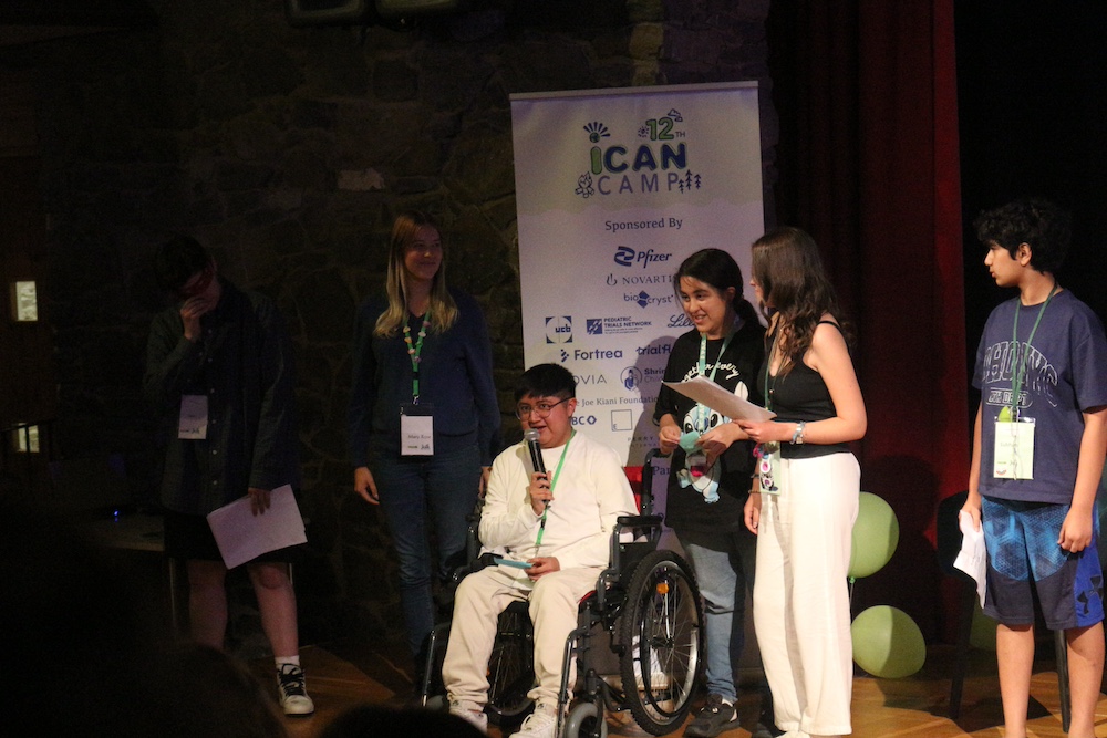 Un beneficiario de APAC y miembro de KIDS Mexico City, usuario de silla de ruedas, presenta con micrófono en el escenario de la Cumbre iCAN 2026, acompañado por jóvenes de otros capítulos de iCAN alrededor del mundo.